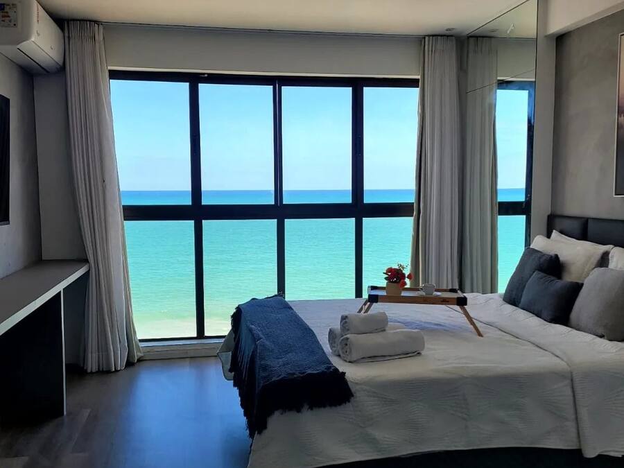 A hotel room with a large window offering a view of the sea, a bed with pillows and a blanket, a bedside table with a tray, and an air conditioning unit.
