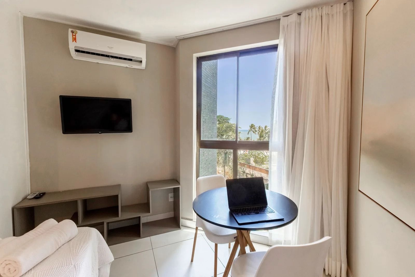 A hotel room with a bed, a TV mounted on the wall, a laptop on a small table, and a window with curtains.