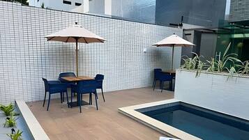Zona junto a la piscina con mesa y sillas, sombrillas y una piscina pequeña.