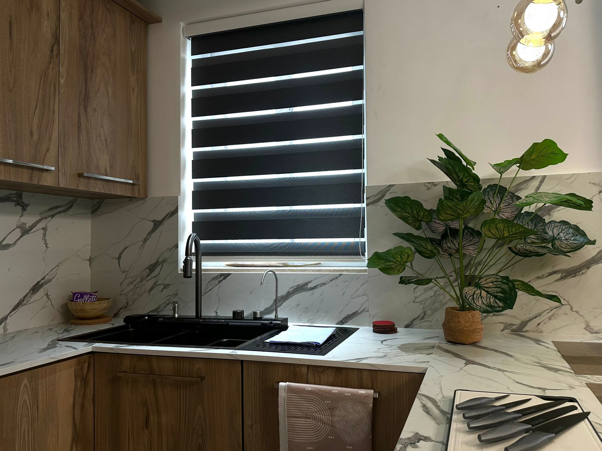 Una cocina con un lavamanos, armarios de madera, salpicadero de mármol, una ventana con persianas y una planta en maceta.