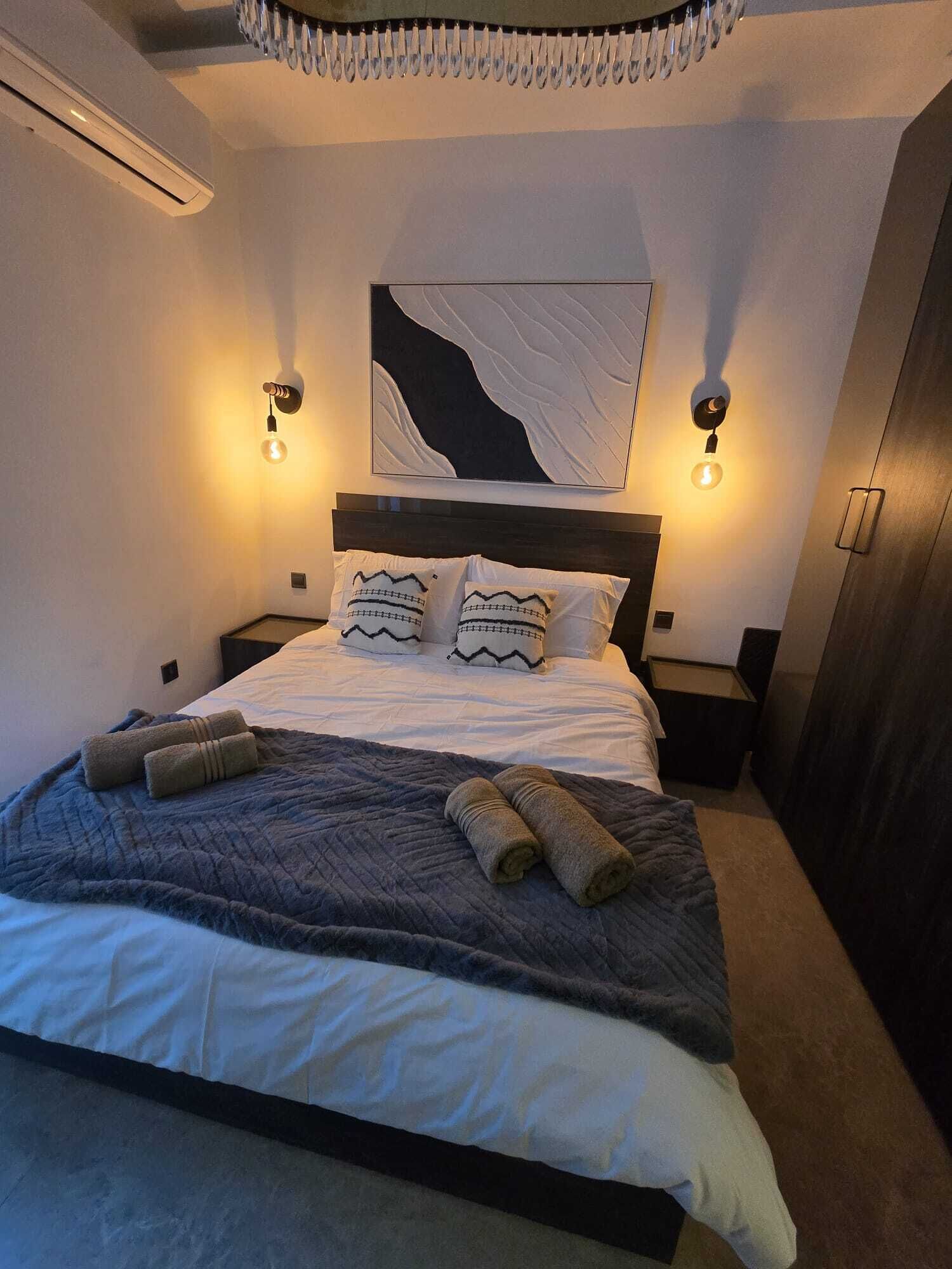 Ein ordentlich gemachtes Bett mit einer gesteppten Decke, zwei Kissen mit Zickzackmuster und aufgerollten Handtüchern. Darüber befindet sich ein an der Wand befestigtes Kunstwerk und zwei Wandleuchten mit freiliegenden Glühbirnen.