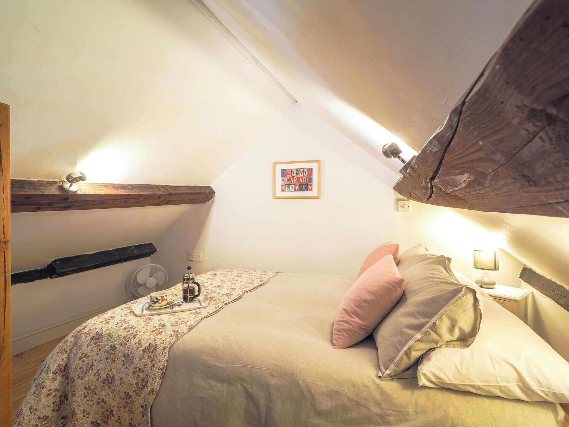 A cozy attic bedroom with a sloped ceiling, a wooden beam, a framed picture, a bedside table with a lamp, and a bed with pillows.