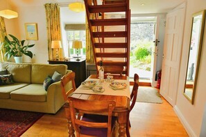 A cozy living room with a wooden dining table set for four, a sofa, a staircase, and a view of greenery outside. - The Cottage at Dolau Afon, Aberystwyth (Aberystwyth)
