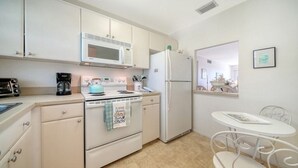 A compact kitchen with a microwave, stove, refrigerator, and a small dining area with a table and chairs. - Siesta Key Beach Unit 306 (Sarasota)