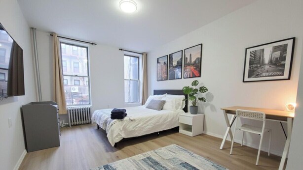 A bedroom with a bed, bedside table, chair, radiator, window, and a desk with a plant. - Simple, Quiet & Clean w/ Workstations (New York)