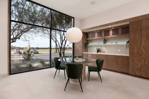A modern dining area with a round glass table, green chairs, a wooden cabinet, and a large window with a view of trees and a distant building.