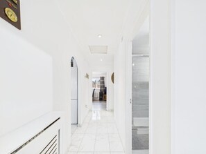 Een gang met witte wanden en een betegelde vloer, die leidt naar een kamer met een bed en een raam.