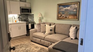 A living room with a sofa, a painting, and a kitchen area with cabinets and appliances. - Tranquil 1-bedroom condo with AC in Madeira Beach completely updated . (Madeira Beach)