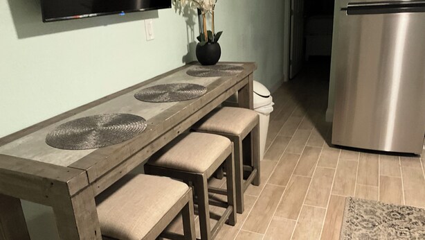 A kitchen with a wooden table, three stools, a vase with flowers, a TV mounted on the wall, and a stainless steel refrigerator. - Tranquil 1-bedroom condo with AC in Madeira Beach completely updated . (Madeira Beach)