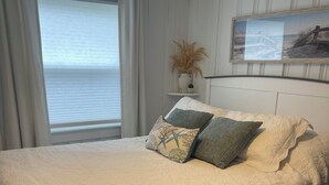 A bedroom with a bed, a window with curtains, a small table with a plant, and a framed picture on the wall. - Cozy 2-bedroom condo located directly on Hampton Beach with AC (Hampton)