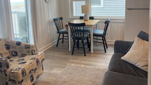 A living room with a gray sofa, a patterned armchair, a dining area with a table and chairs, and a kitchen with a refrigerator. - Cozy 2-bedroom condo located directly on Hampton Beach with AC (Hampton)