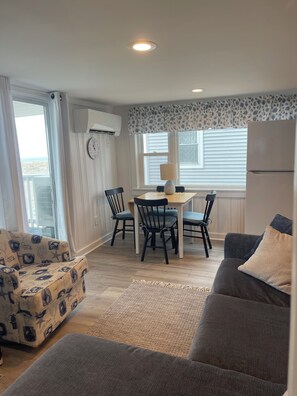 Ein Wohnzimmer mit grauem Sofa, einem gemusterten Sessel, einem Essbereich mit Tisch und Stühlen sowie einer Küche mit Kühlschrank.