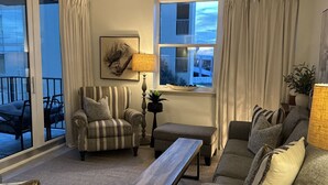 Un salon comprenant un canapé, un fauteuil et une table basse en bois. Il y a une fenêtre avec des rideaux, une lampe et un tableau encadré au mur.