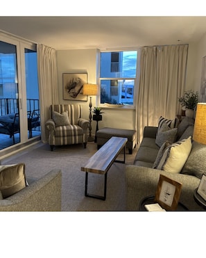 Un salon comprenant un canapé, un fauteuil et une table basse en bois. Il y a une fenêtre avec des rideaux, une lampe et un tableau encadré au mur.