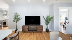 A modern living room with a flat-screen TV, wooden flooring, and a dining area with a white table and chairs. - Stunning 4BR2BA w Parking/Near Downtown/ +Parking (Boston)