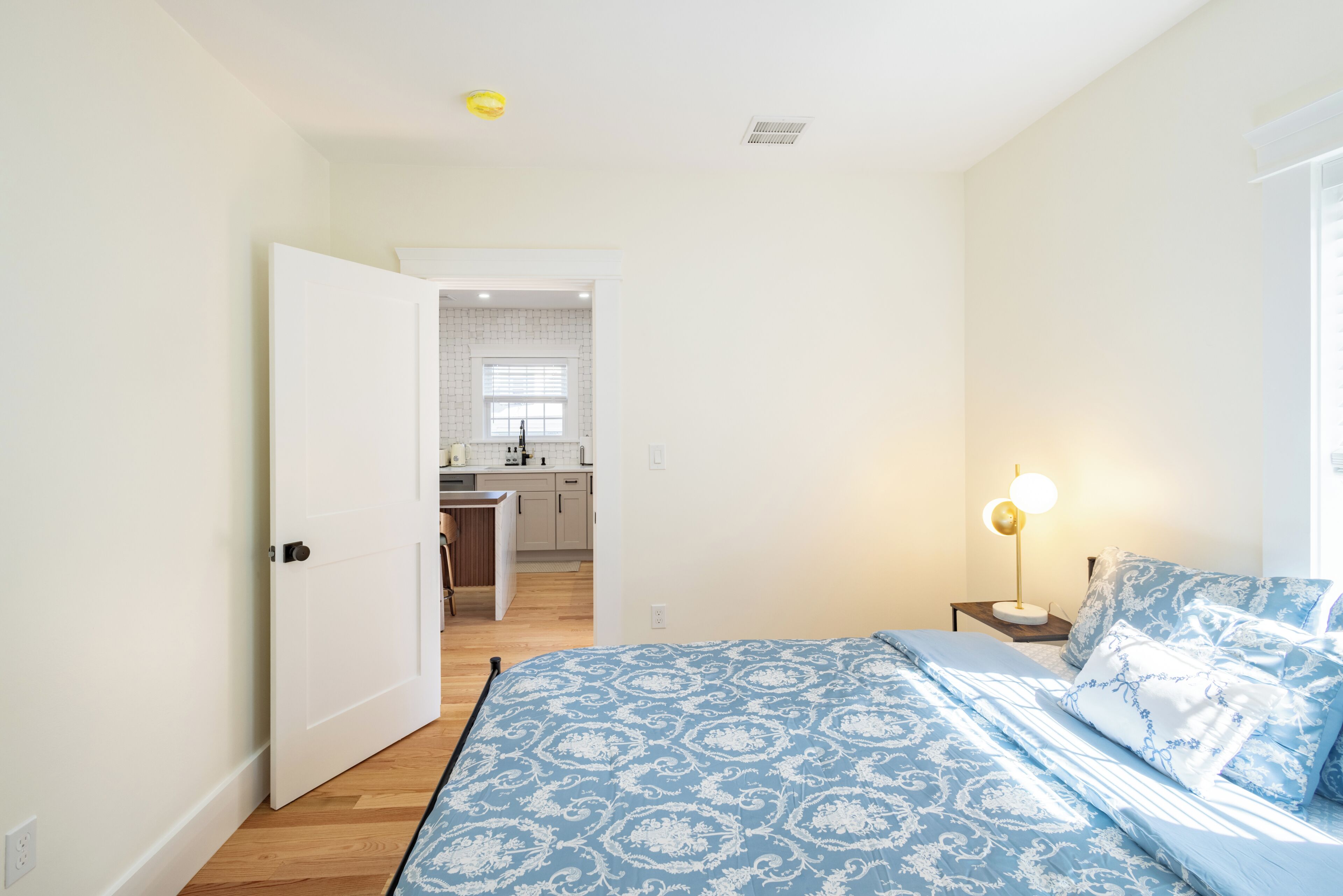 A bedroom with a patterned bedspread, a bedside lamp, and a view into the kitchen.