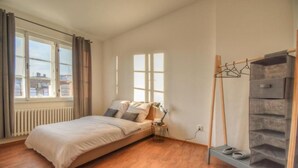 A bedroom with a bed, a radiator, a window with curtains, a shoe rack, and a lamp.
