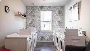 A laundry room with two washing machines, a red stool, a window, and a wall-mounted shelf with jars.