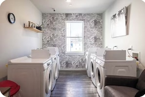 A laundry room with two washing machines, a red stool, a window, and a wall-mounted shelf with jars.