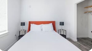 A hotel room with a large bed, two bedside tables with lamps, a window with blinds, and a door leading to a hallway.