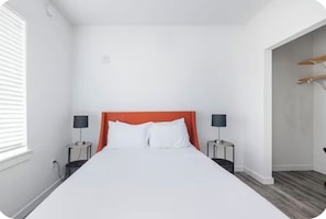 A hotel room with a large bed, two bedside tables with lamps, a window with blinds, and a door leading to a hallway.