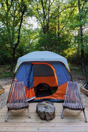 Una carpa con entrada abierta, dos sillas de madera y un tronco sobre una terraza de madera en un bosque.