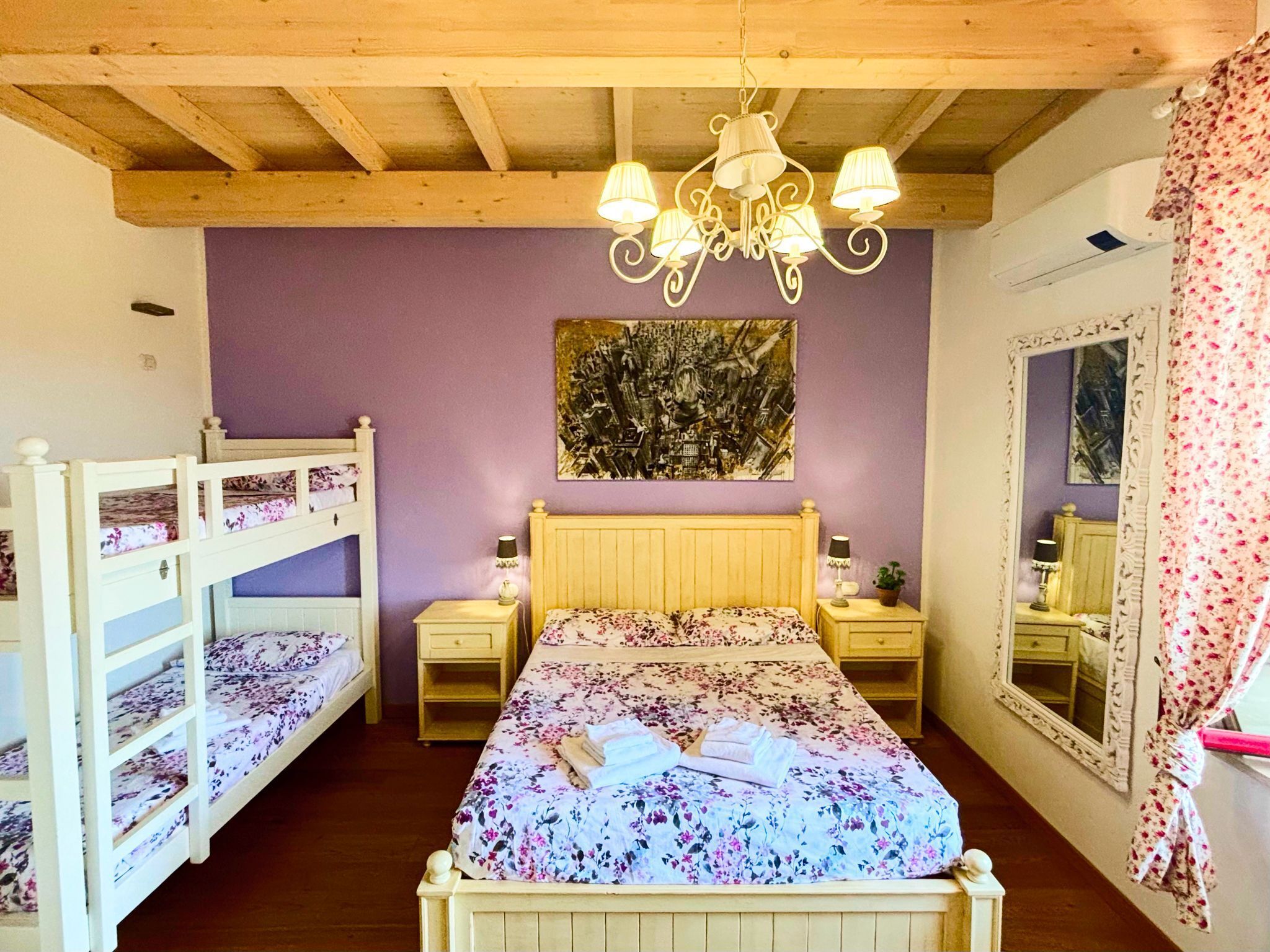 A bedroom with a bunk bed, a single bed, a chandelier, bedside tables, a mirror, and a painting on the wall.