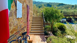 A stone building with a staircase leading to a terrace, flags, and a view of a parking area and hills. - NonnaNara (Grosseto)