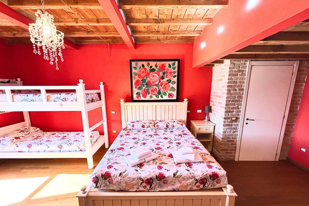 A bedroom with a wooden bunk bed, a single bed with floral bedding, a chandelier, a framed floral painting, a nightstand, and a brick wall.