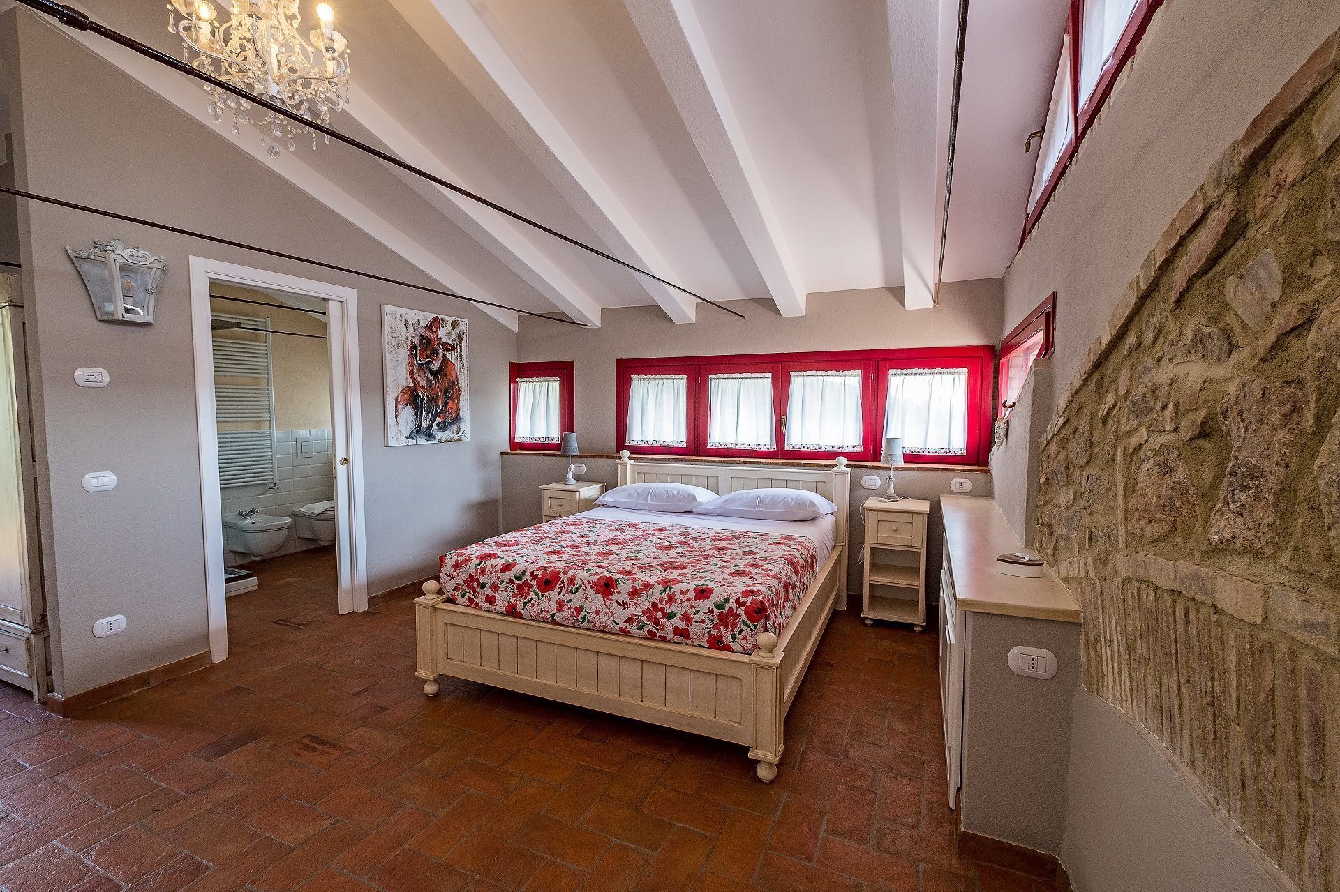Un dormitorio con cama, mesitas de noche, ventana con marcos rojos y un candelabro.