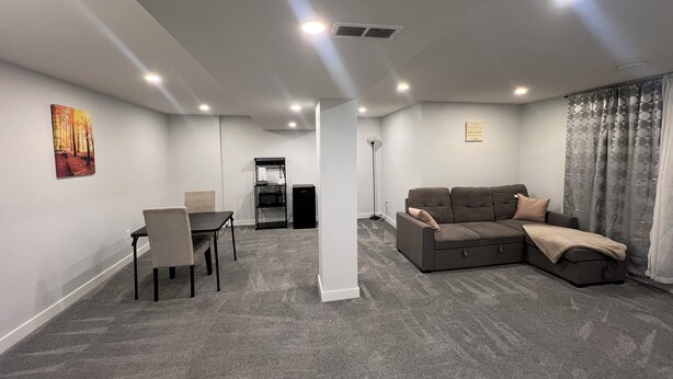 A modern living room with a sectional sofa, a dining table with chairs, a small black cabinet, and a standing floor lamp. - Hidden Gem 💎 Stylish Suite w/ All-Inclusive Comfort” (Regina)