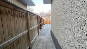 A narrow concrete pathway flanked by a wooden fence and a textured wall, leading to a wooden deck. - Hidden Gem 💎 Stylish Suite w/ All-Inclusive Comfort” (Regina)