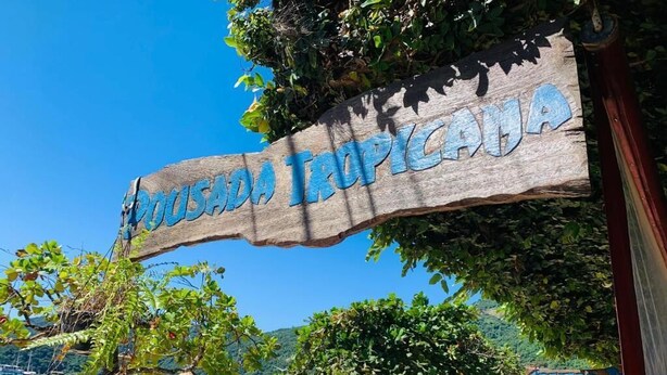 A wooden sign with 'TROPICALA' written on it, a beach, boats, and a clear blue sky. - Pousada Tropicana (Angra dos Reis)