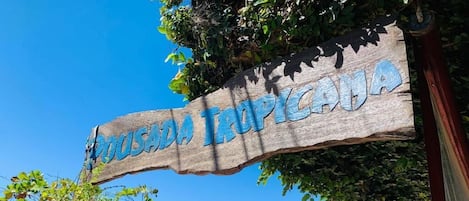 A wooden sign with 'TROPICALA' written on it, a beach, boats, and a clear blue sky.