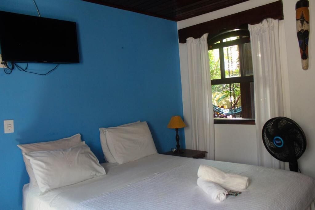 A room with a bed, a TV mounted on the wall, a window with curtains, a lamp, and a fan.