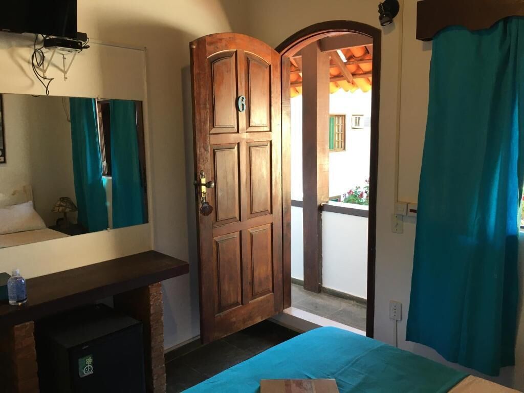 A room with a wooden door, a bed with a blue bedspread, a small table with a bottle, and a mirror.