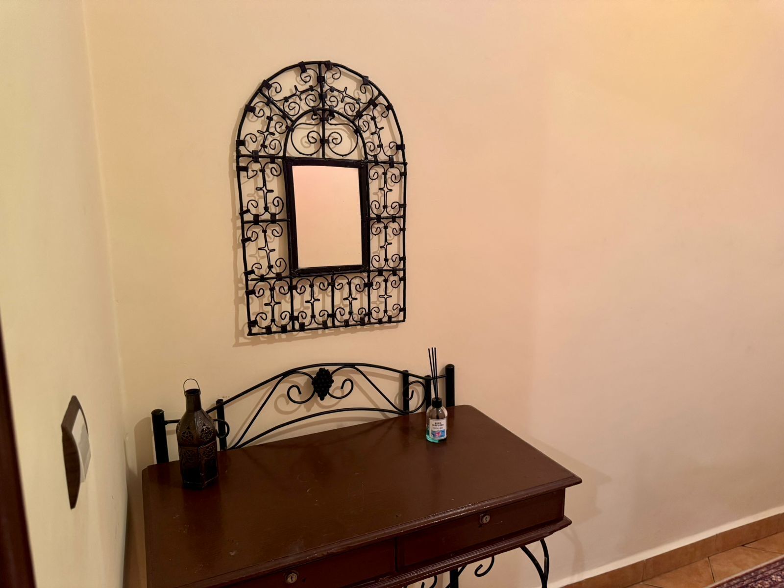 Una habitación con una pequeña mesa de madera, un espejo metálico decorativo y una botella de gel hidroalcohólico.