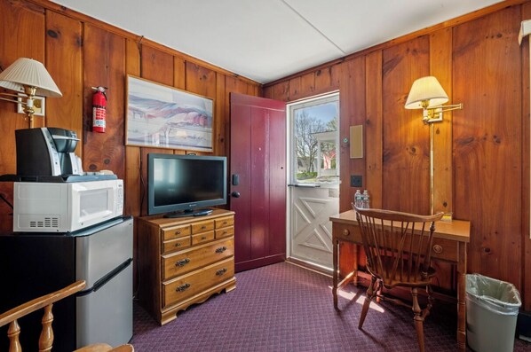 A room with wooden walls, a television on a wooden dresser, a microwave, a coffee maker, a lamp, a chair, and a door leading outside.