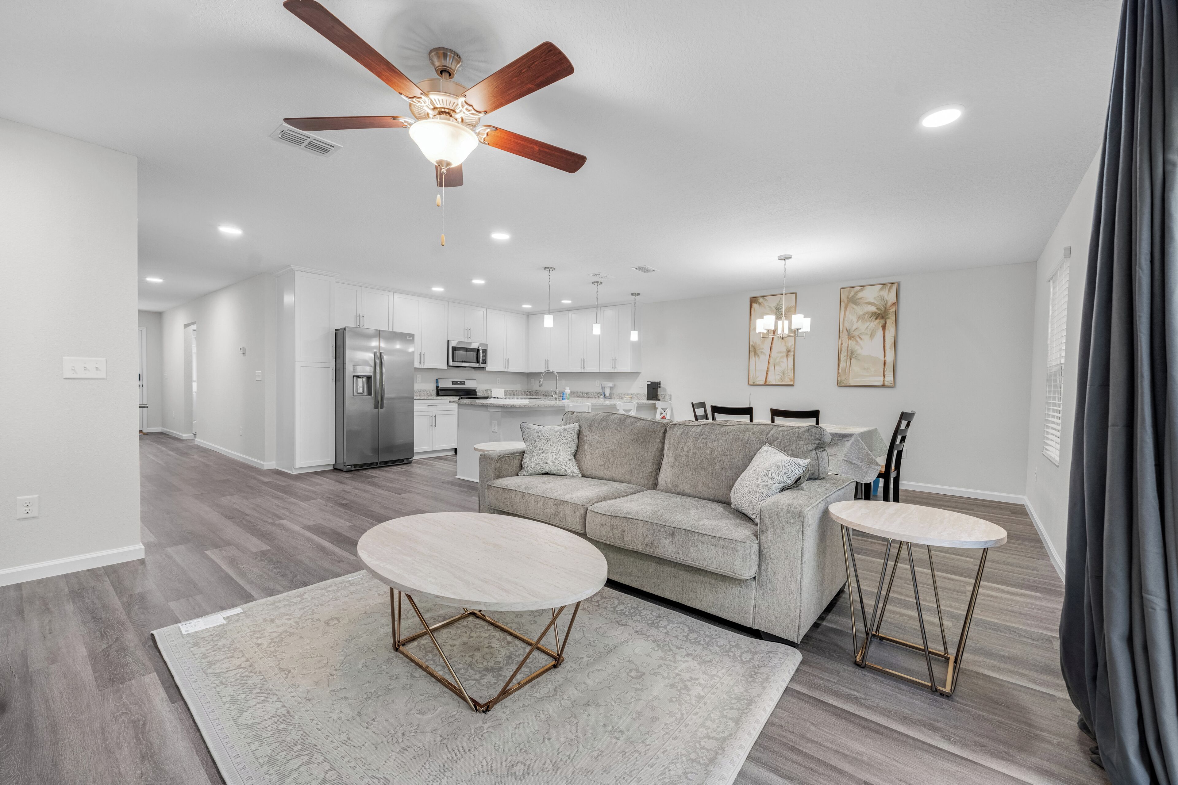 A modern living room with a sofa, coffee table, and dining table, connected to a kitchen area with a ceiling fan and recessed lighting.