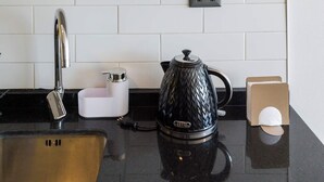 Een keuken met een zwarte waterkoker, een koffiezetapparaat en een spoelbak. Boven de spoelbak zijn planken met drinkglazen en potten.