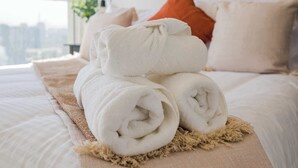 Een net opgemaakt bed met opgerolde handdoeken, een beige plaid en een stadszicht door een groot raam.