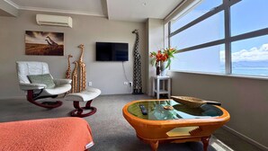 Una habitación con un ventanal grande, televisión, una escultura de jirafa, una mecedora y una mesa de centro de vidrio.