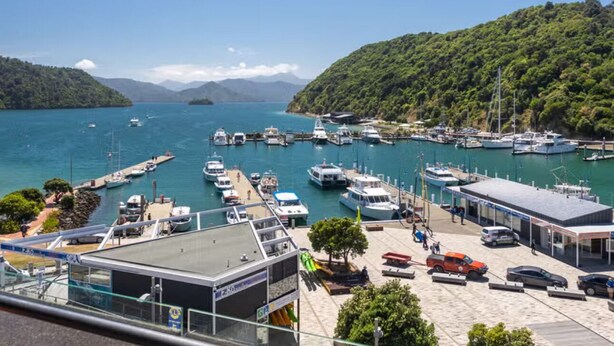 Un puerto deportivo con barcos amarrados, un edificio de techo plano y un paisaje montañoso al fondo.