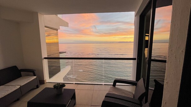 A balcony with a view of the sea at sunset, a sofa, a small table, and a potted plant. - Luxury Beachfront Apartment with Panoramic Ocean Views – Strand Ocean View 1004 (Strand)