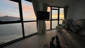 A room with a large window overlooking a body of water, a flat-screen TV, a sofa, and a pair of legs. - Luxury Beachfront Apartment with Panoramic Ocean Views – Strand Ocean View 1004 (Strand)