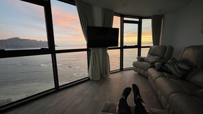 Ein Zimmer mit großem Fenster mit Blick auf ein Gewässer, einem Flachbildfernseher, einer Couch und einem Paar Beine.