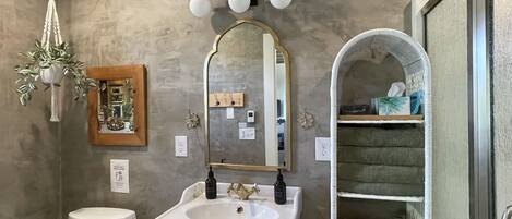 A bathroom with a white sink, a mirror, a hanging plant, and a shelf with toiletries.