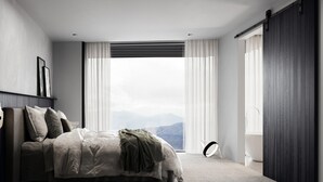 Ein Schlafzimmer mit einem Bett, einem Nachttisch, einem Fenster mit Vorhängen und Blick auf Berge.