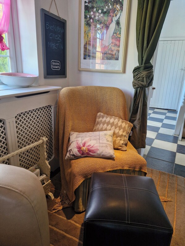 A cozy room with a cushioned chair, a black leather ottoman, a framed artwork, and a chalkboard with a welcome message.