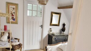 Quarto com cama, penteadeira, cadeira, prateleira com objetos, porta e luminária suspensa.